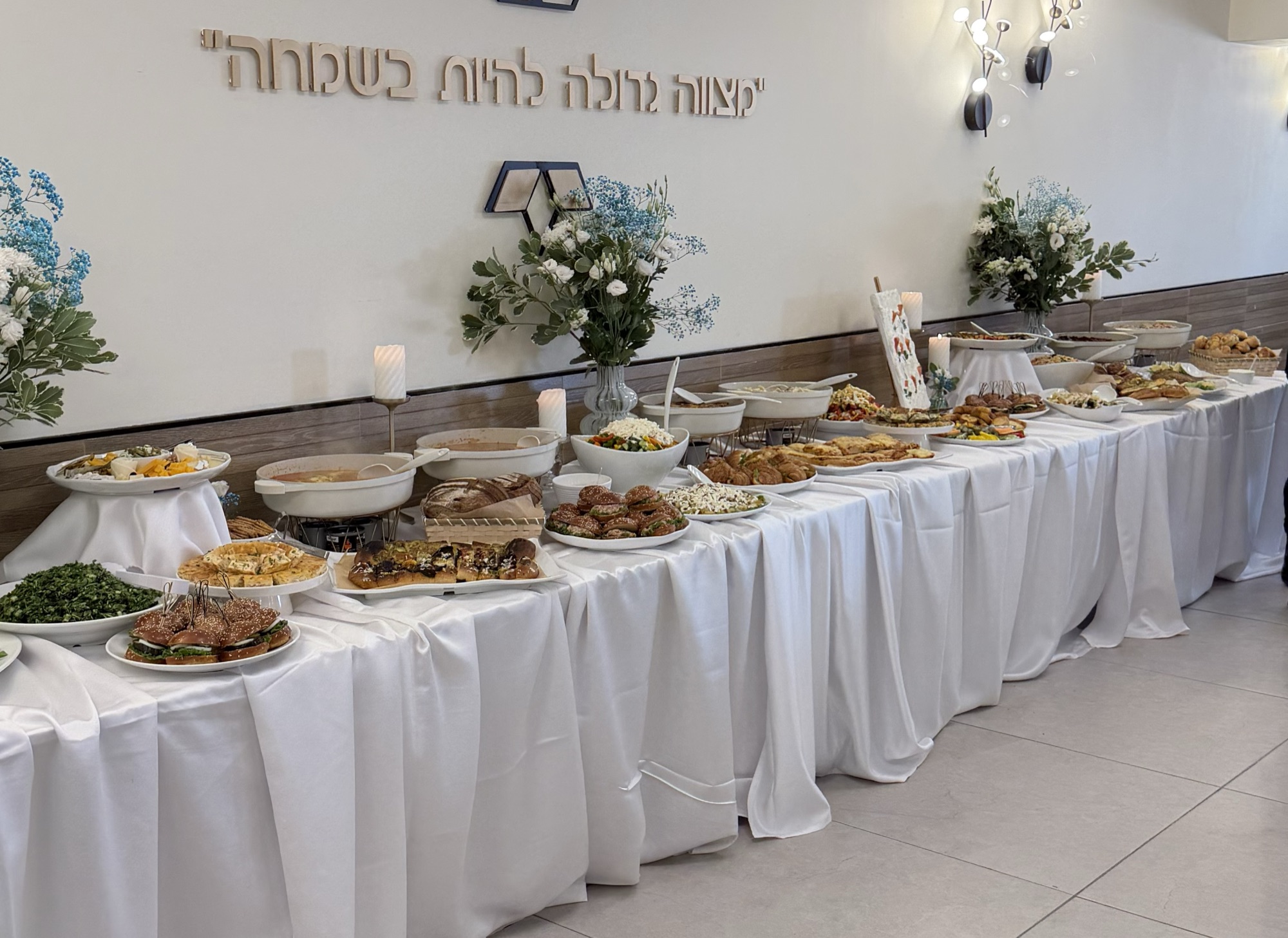 שולחן בופה ארוך ערוך בכלי הגשה לבנים, מפות לבנות, פרחים ונרות